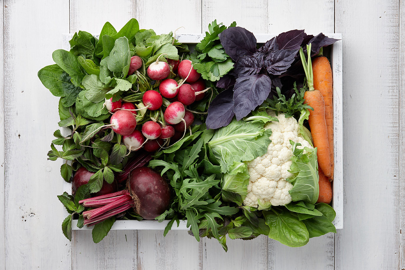 A wooden crate filled with a variety of fresh vegetables, including radishes, carrots, cauliflower, leafy greens, and herbs, arranged on a white wooden surface.