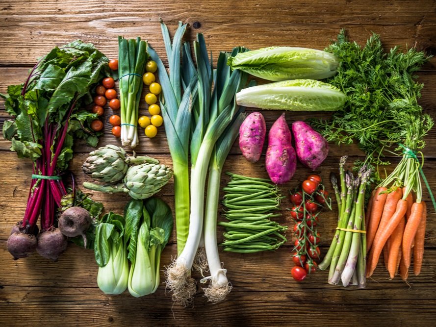 Fresh assortment of colorful vegetables including beets, artichokes, leeks, lettuce, sweet potatoes, carrots, asparagus, green beans, and cherry tomatoes arranged on a wooden table. Ideal for healthy eating and cooking inspiration.