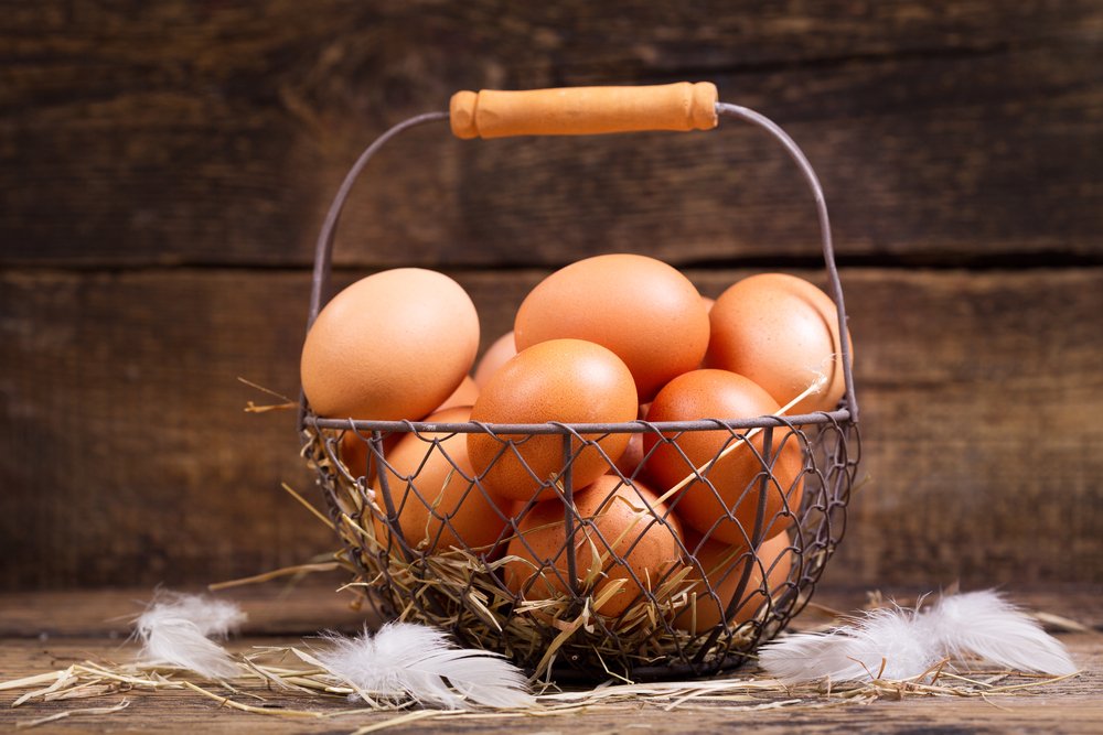 Basket of fresh brown eggs resting on straw with soft white feathers, set against a rustic wooden background. Ideal for farm-to-table themes and culinary content.